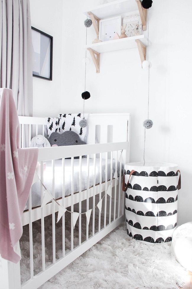 اتاق خواب نوزاد شیک و مدرن که دیوار سفید، تخت نوزاد سفید، پرده خاکستری و ریسه مثلثی و گلوله ای دارد